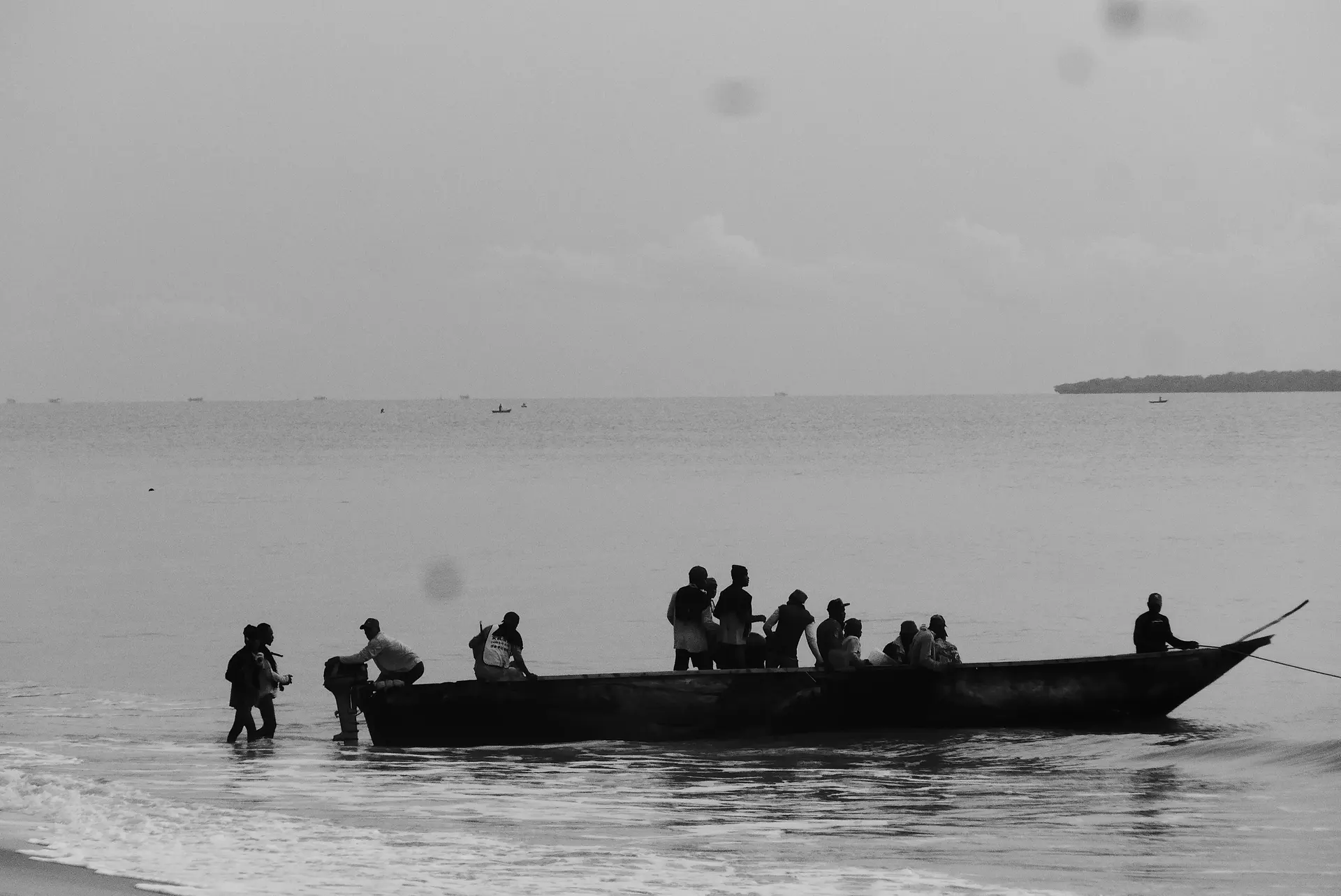 Black-and-white photo of people in a long wooden motorboat in shallow surf, an island on the horizon