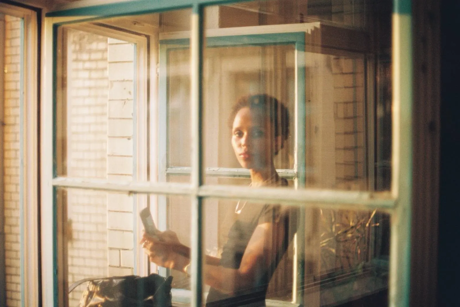 Woman holding a phone, seen through a green window frame in warm sunlight